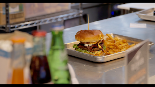 Close-up of stacked cheeseburger with bacon and lettuce served with fries on tray amid bottles – appetizing restaurant promotional videos footage for food hall menu showcases