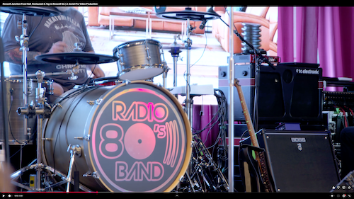 Close-up of neon 'Radio 80's Band' drums with drummer and amps on purple-curtained stage – dynamic venue promo video campaigns footage for Roswell Junction events